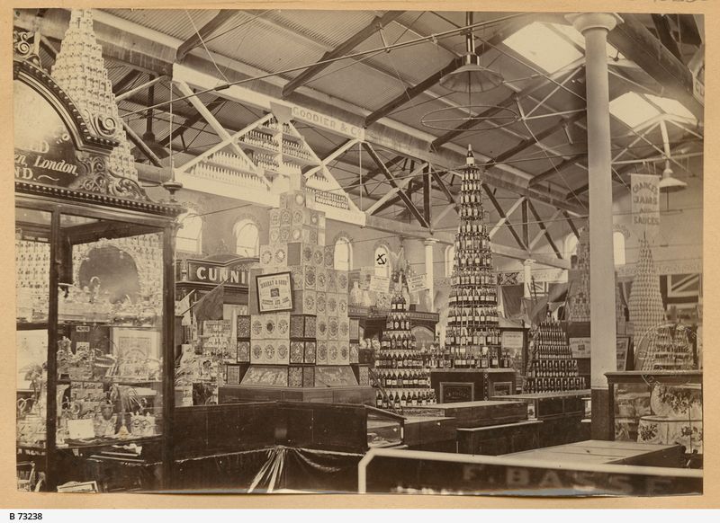 Sepia photograph of a large exhibition hall interior with high exposed iron roof trusses and skylights, showing several tall pyramid-shaped displays built from stacked tins and jars, trade signs for various manufacturers on boards and banners, and iron columns running the length of the hall