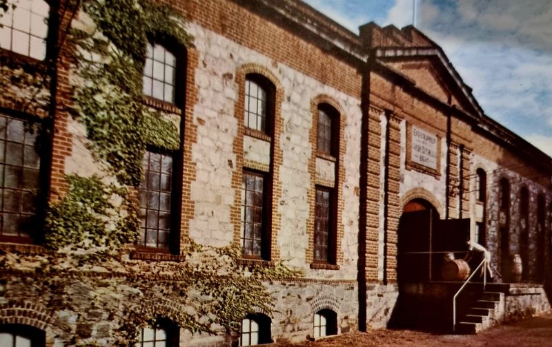 Colourised photograph of the Chateau Reynella cellar block — a long two-storey stone and brick building with arched windows covered in ivy, a wine barrel on a trolley beside the entrance steps