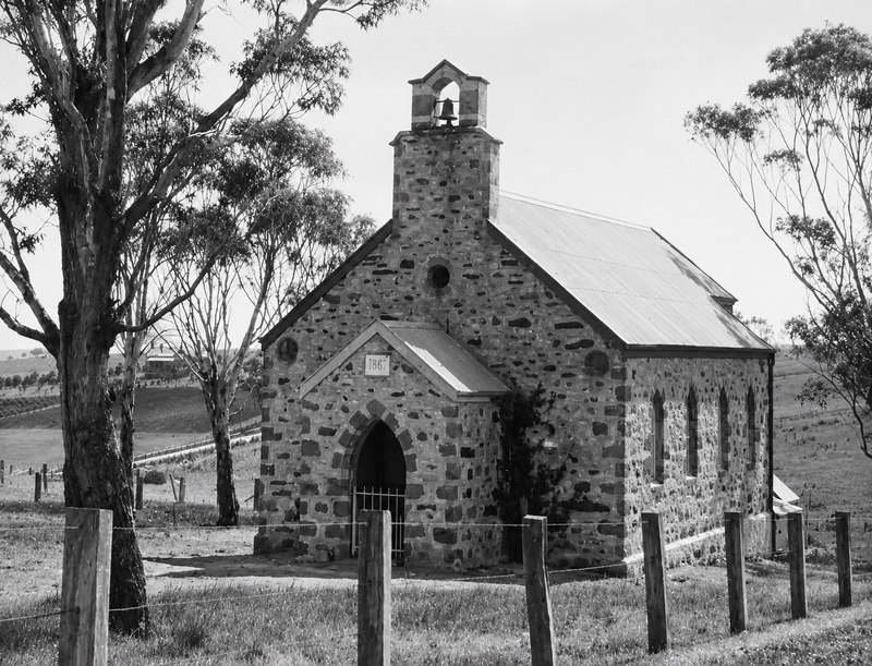 Black-and-white photograph of a small rubble-stone church with a bellcote, arched doorway, and corrugated iron roof, surrounded by a wire fence with gum trees behind on a rural hillside