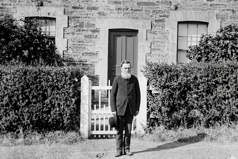 Black-and-white photograph of a man in dark clothing standing before a stone building, a small white gate beside him, plants flanking the wooden doorway