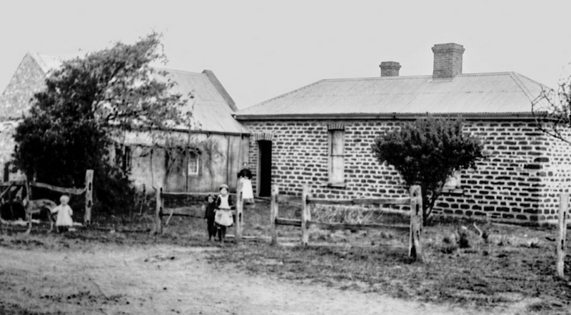 Black-and-white photograph of a low stone and brick farmhouse with a chimney, several figures standing before it, young trees in the foreground and open cleared land around