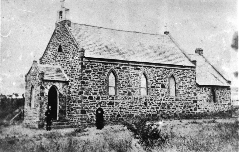 Black-and-white photograph of the original St Mary's Catholic Church — a rough-cut stone building with Gothic pointed arched windows, a slate roof, bellcote, and two figures at the arched entrance