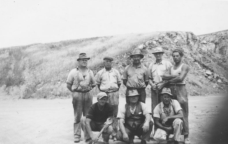 Black-and-white photograph of eight quarry workers — five standing and three crouching — posing in front of a steep quarry face at Reynella, 1940