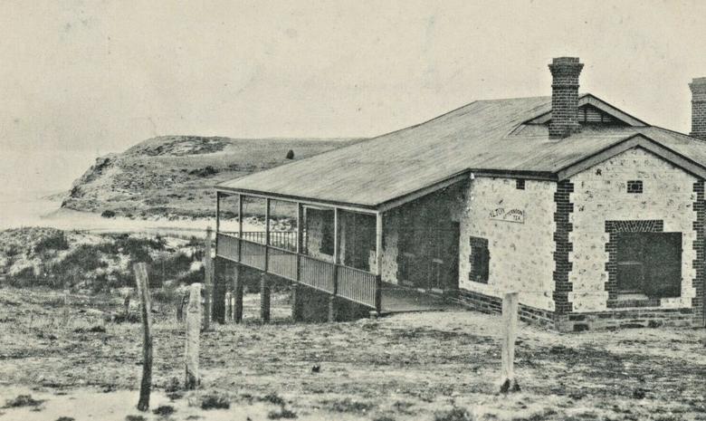 Black-and-white photograph of a low single-storey stone building with a corrugated iron roof, open timber verandah and chimney, set in bare sandy dunes with clifftops visible in the background to the left
