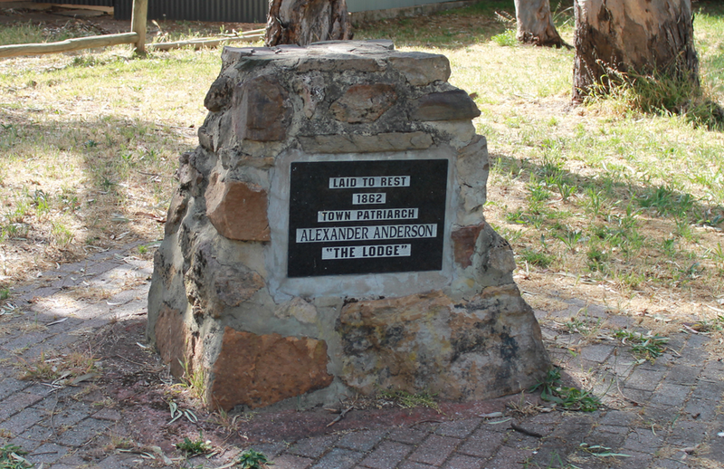 Rough stone cairn with a rectangular dark metal plaque mounted on its face, standing on a paved path beneath gum trees at Creighton Avenue, Morphett Vale — the burial marker for Alexander Anderson