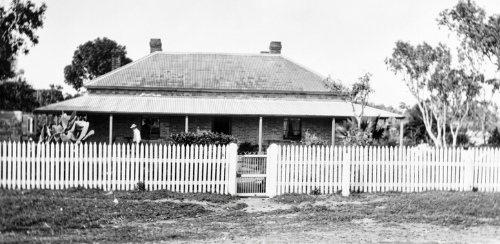 Black-and-white photograph of a low single-storey homestead with a hipped roof behind a white picket fence, a figure on the verandah and gum trees behind