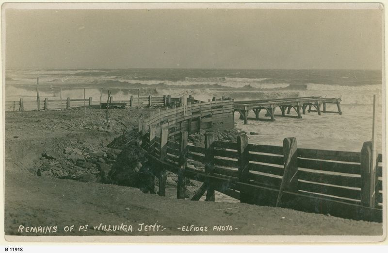 Sepia postcard photograph of the storm-damaged Port Willunga timber jetty with sections of decking and piling collapsed and skewed, heavy surf breaking in the background