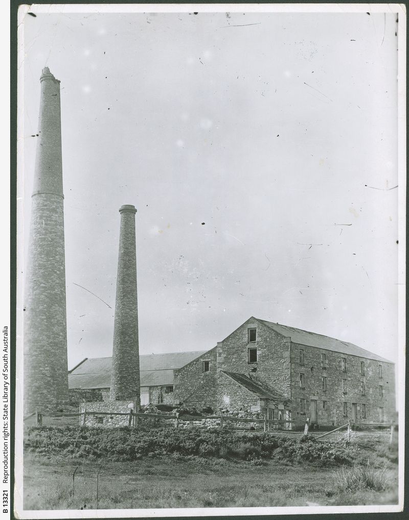 Black-and-white photograph of White's Flour Mill at Aldinga — two tall brick chimney stacks dominating the left side, with a multi-storey stone mill building behind a fence
