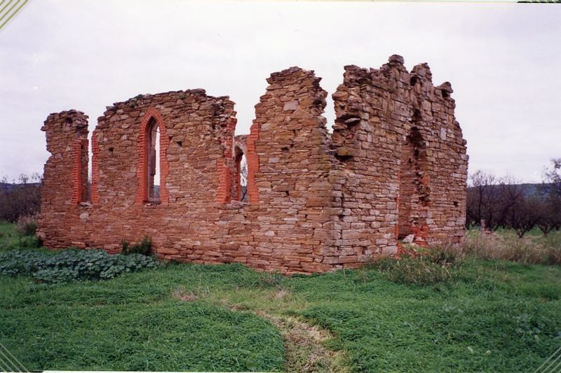 Roofless ruins of a stone church, the walls of warm ochre rubble with red brick dressings framing tall pointed Gothic-arched window openings; green grass in the foreground and a row of trees behind under an overcast sky