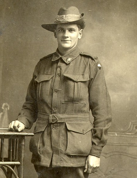 Black-and-white studio portrait of a young man in World War I Australian military uniform, wearing a slouch hat and khaki jacket, standing beside a small table