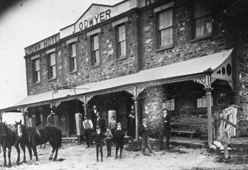 Black-and-white photograph of a two-storey rough-stone hotel with a covered ground-floor verandah, decorative timber posts, a sign above the verandah, horses tethered out front and men standing on the verandah