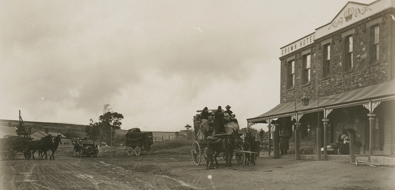 Black-and-white photograph of Old South Road, Reynella, with a horse-drawn vehicle on the unpaved road in the foreground and the two-storey Crown Hotel visible on the right, open paddocks and gum trees in the distance