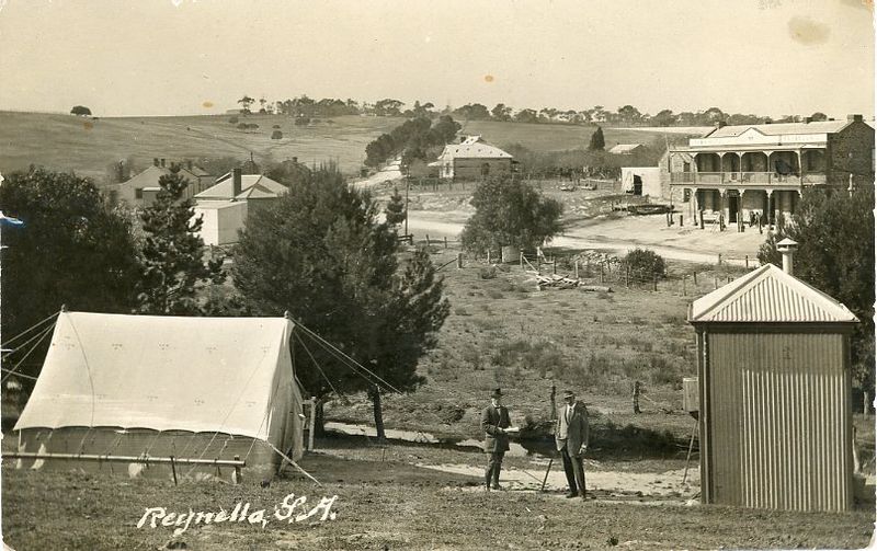 Panoramic black-and-white photograph looking down over Reynella township, with a white canvas tent in the left foreground, two men conversing on a path, a corrugated iron shed at lower right, and the Crown Hotel with its upper balcony visible among trees in the middle-right background