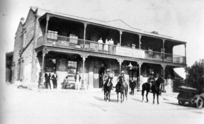 Black-and-white photograph of the two-storey Crown Hotel with an ornate upper balcony featuring decorative ironwork, women standing on the balcony, men and horses on the road in front and a motor car to the right