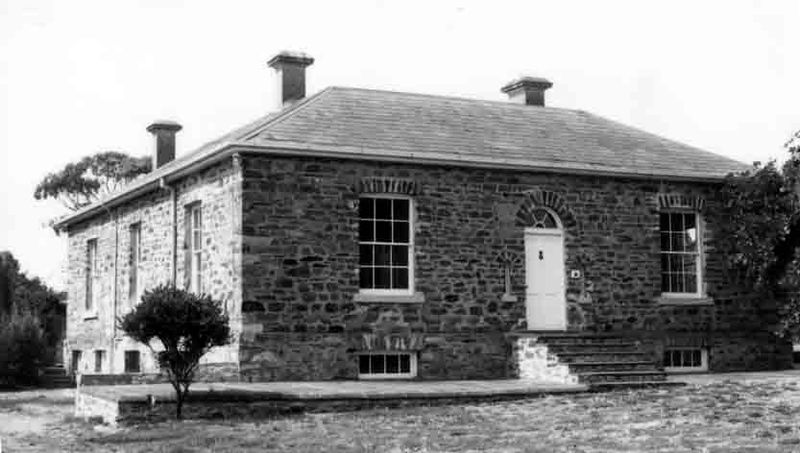 Black-and-white photograph of a solid two-storey rubble stone building with a hipped slate roof and three chimneys, large sash windows with brick arch dressings, a central front door with a fanlight above stone steps, and basement windows set low in the front wall, with a small clipped shrub at the front left and trees visible behind