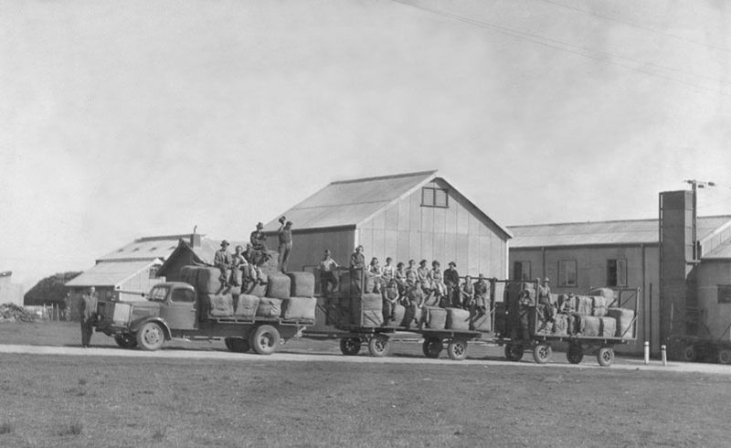 Black-and-white photograph of a truck and trailer piled with bales of flax, workers sitting on top of the load outside large corrugated iron shed buildings at the Morphett Vale Flax Mill