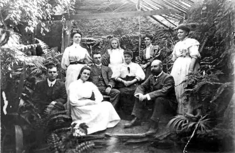 Black-and-white family portrait of nine people posed in a conservatory surrounded by tropical ferns and plants; a bearded man is seated at right and a woman seated centrally, with younger men, women, and a girl arranged around them in formal late-Victorian dress