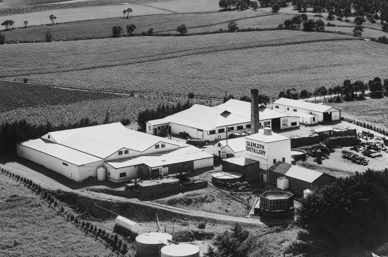 Aerial black-and-white photograph of the Glenloth Winery complex — corrugated iron sheds, a tall chimney stack lettered Glenloth Distillery, storage tanks, and surrounding vineyard paddocks