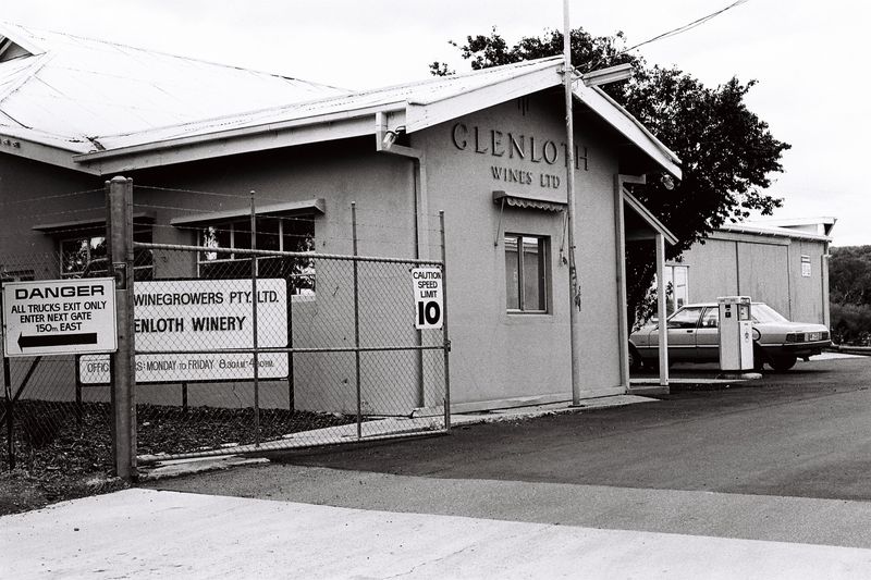 Black-and-white photograph of the Glenloth Wines Ltd entrance — a rendered office with Glenloth Wines Ltd on the facade, chain-link fence with winery signage, and a car beside the building