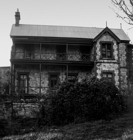 Black-and-white photograph of a two-storey stone house with a verandah and iron balcony railing on the upper floor, Gothic-arched windows, and an overgrown garden in the foreground