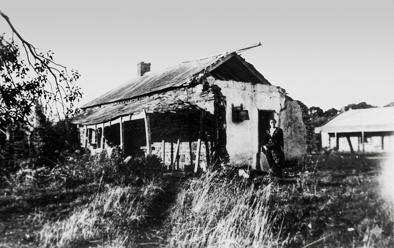 Black-and-white photograph of a derelict wattle-and-daub building with a badly sagging corrugated iron roof, a woman standing at the entrance, long grass in the foreground
