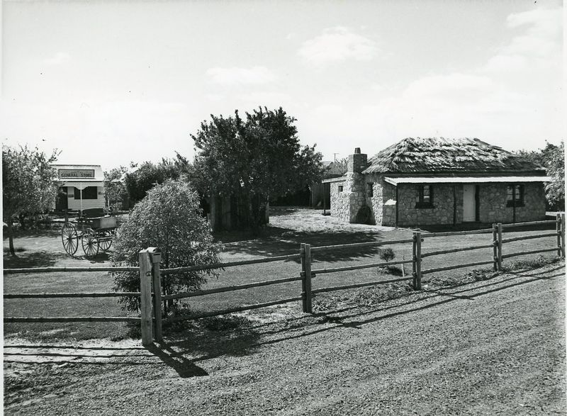 Black-and-white photograph of a low stone building with a thatched roof behind a wooden post-and-rail fence, a replica general store with a horse-drawn cart to the left, trees in the background
