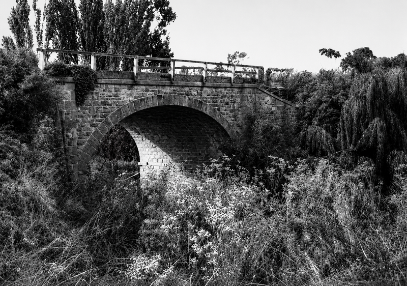 Black-and-white photograph of a stone arched bridge over an overgrown creek — a single arch of coursed stone and brick with a cast iron railing, surrounded by poplars and scrub