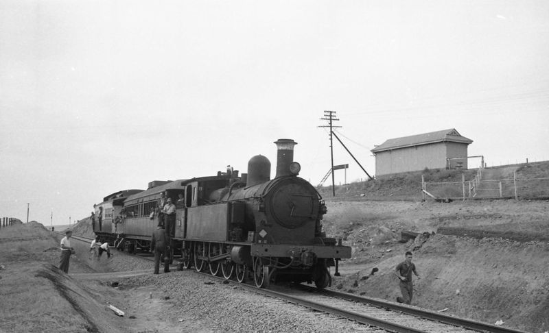 Black-and-white photograph of a steam locomotive with passenger carriages stopped at a cutting, passengers beside the track and a small station building above on the right