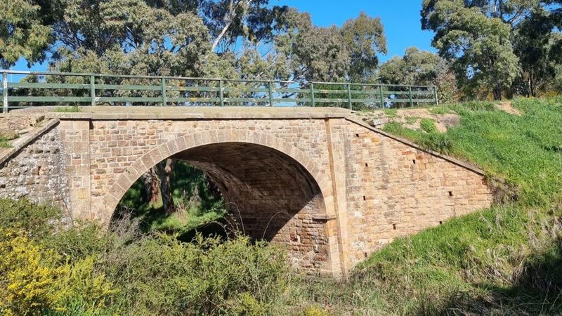 Colour photograph of Hallett's Bridge — a single-arch stone bridge of coursed sandstone with a metal railing, surrounded by green grass, scrub, and gum trees on a clear day