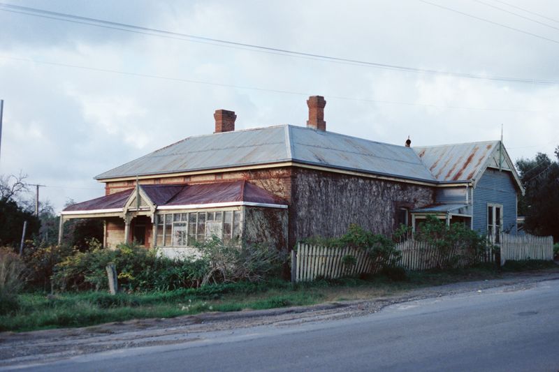 Colour photograph of a single-storey limestone cottage with a corrugated-iron hipped roof and two brick chimneys, a verandah across the front with a central gabled entrance and one end enclosed with windows, a timber-clad gabled extension at the right, a picket fence and overgrown garden in front, beside a sealed road under a cloudy sky