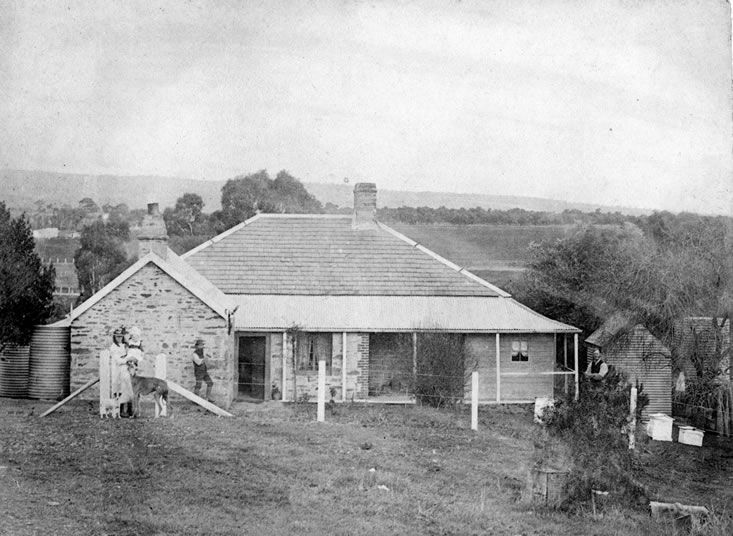 Black-and-white photograph of a single-storey stone farmhouse with a hipped corrugated-iron roof, a verandah and a brick chimney, a gabled stone wing to the left with its own chimney, round corrugated water tanks beside it, a woman and a man standing in the bare front yard with a dog, beehive boxes at right, and open paddocks and a low range of hills behind