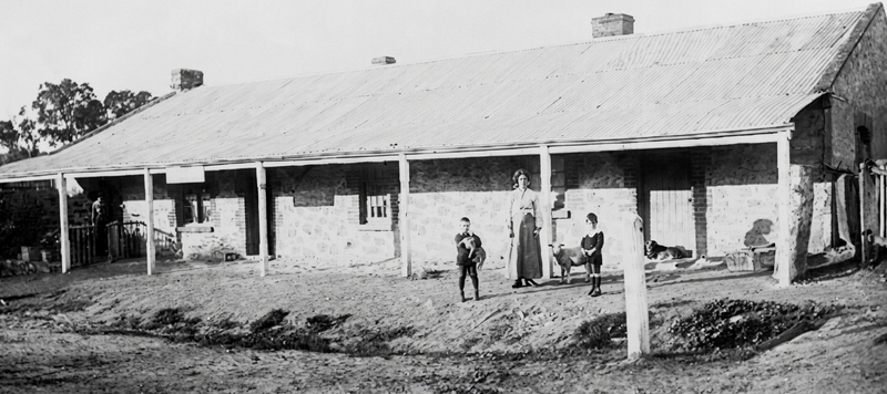 Black-and-white photograph of a long single-storey stone building with a corrugated-iron roof and a timber-posted verandah running its full length; three people stand on the bare ground in front — a man, a woman in a long dark dress, and a child — with household items and outbuildings to either side