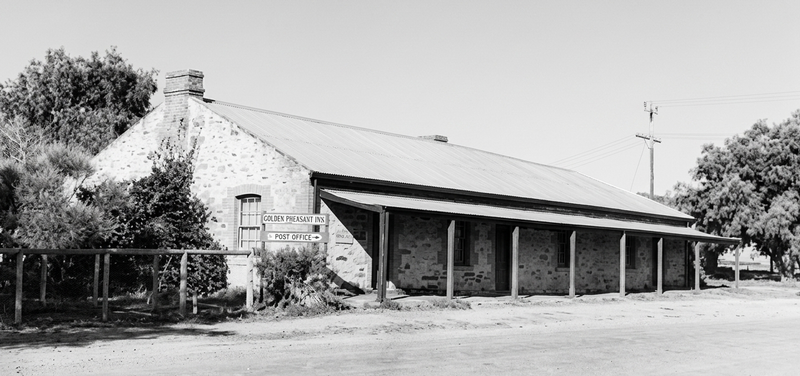 Black-and-white photograph of a long single-storey stone colonial building with a corrugated iron roof and a verandah supported by timber posts, beside a dirt road