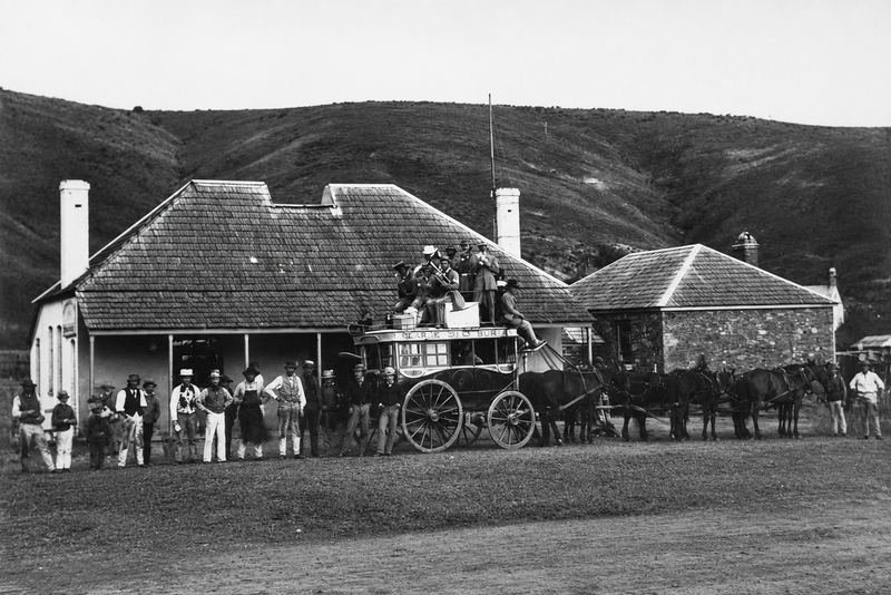 Black-and-white photograph of a horse-drawn coach with luggage on top and a large group of men gathered outside a low stone inn with a tiled hipped roof, the Onkaparinga hills behind