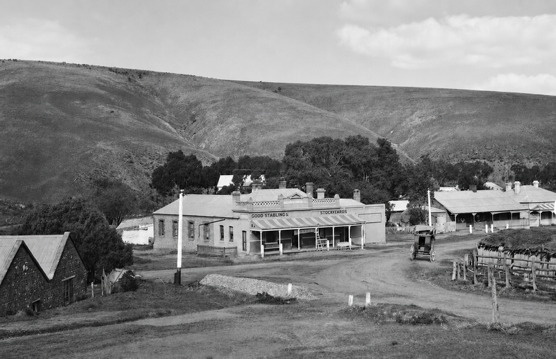 Black-and-white photograph looking down on Noarlunga township — a low building with verandah and stockyard signage on the central block, other buildings on a dirt road, the hills behind