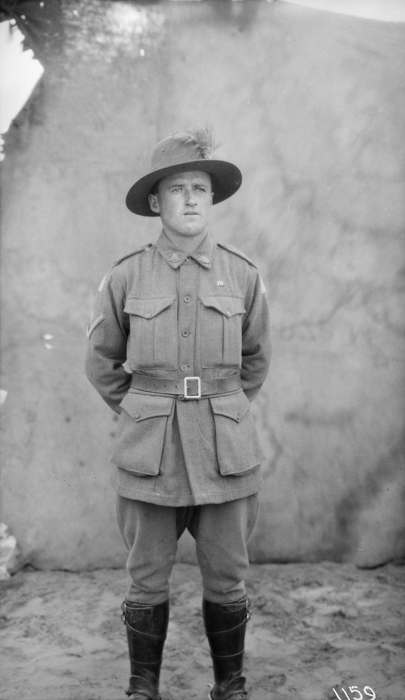 Black-and-white full-length studio portrait of a young soldier standing at ease with his hands behind his back, wearing a slouch hat with a plume, a belted tunic, riding breeches and tall leather boots, against a plain backdrop