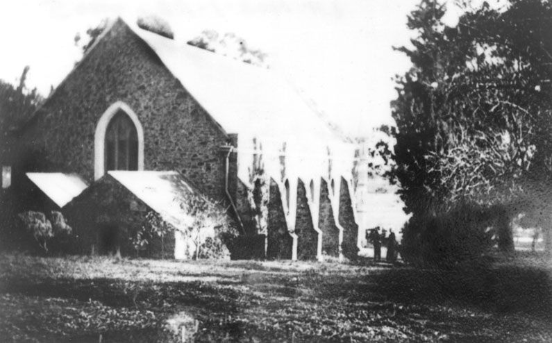 Black-and-white photograph of a stone church with a steep pitched roof and Gothic arched window on the gable, partially obscured by trees; figures in the churchyard