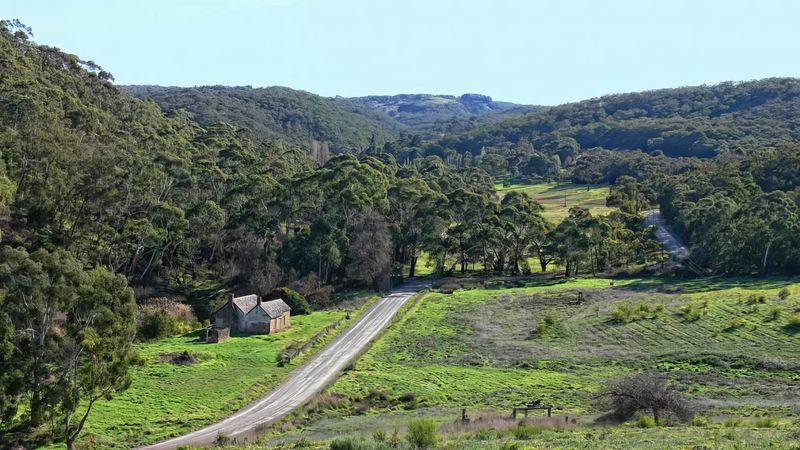 Colour photograph looking down into a wide Adelaide Hills valley with rolling green pasture and eucalyptus forest on the surrounding slopes; a sealed road runs through the centre of the frame, and on the left side in the middle distance stand the roofless stone ruins of the Mackereth Cottage in two small sections, dwarfed by the scale of the surrounding landscape