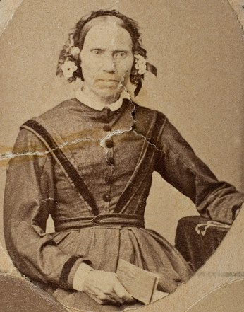 Sepia studio photograph of an older woman seated and facing the camera, wearing a dark high-necked dress with a buttoned bodice and a frilled lace cap tied under the chin with ribbons, her hair parted in the centre, one hand resting beside her and holding a folded white card