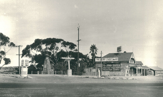 Black-and-white photograph of a rural crossroads with a stone memorial column in the left foreground and a single-storey timber store building with a verandah to the right, large gum trees behind, 1963