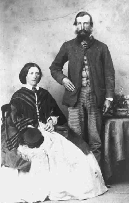 Black-and-white studio portrait of a seated woman in a wide hooped skirt and striped shawl with centre-parted dark hair, a small hat resting in her lap, beside a standing bearded man in a dark jacket and checked trousers with one hand on a draped table
