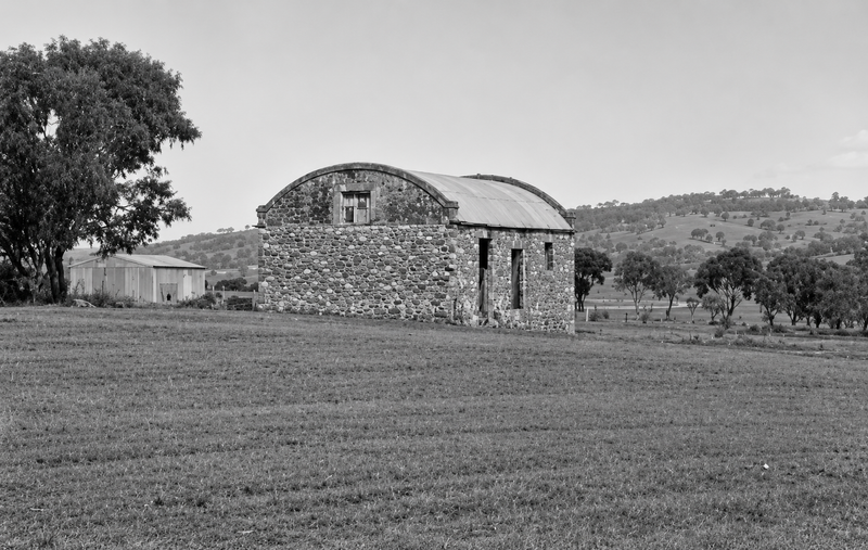 Black-and-white photograph of a large stone barn with a distinctive barrel-vaulted corrugated iron roof, standing in an open green paddock with wooded hills behind