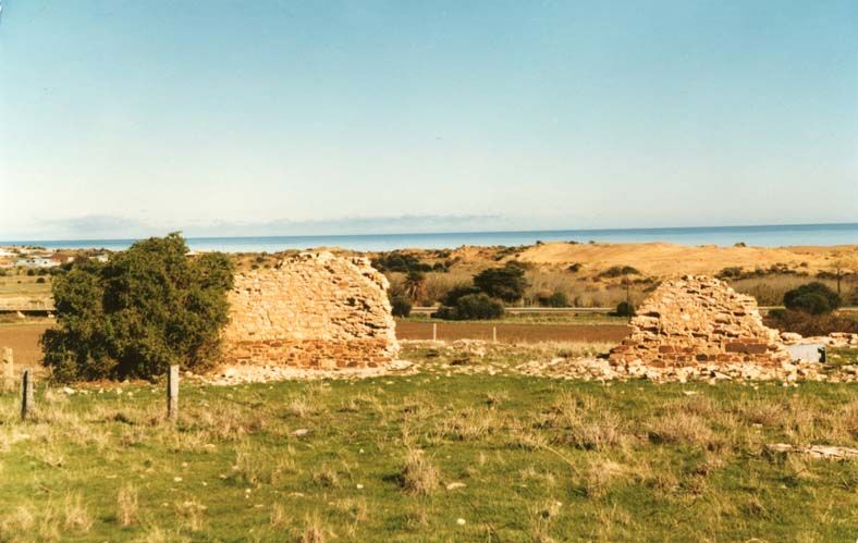 Colour photograph of two low stone gable-end walls — all that remains of the Dalkeith farm buildings — standing on a green coastal clifftop with the Gulf St Vincent visible beyond