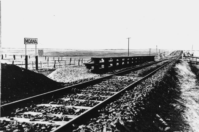 Black-and-white photograph showing the Moana station stop — a Moana nameboard post, a low timber platform, and a single track stretching across flat open farmland