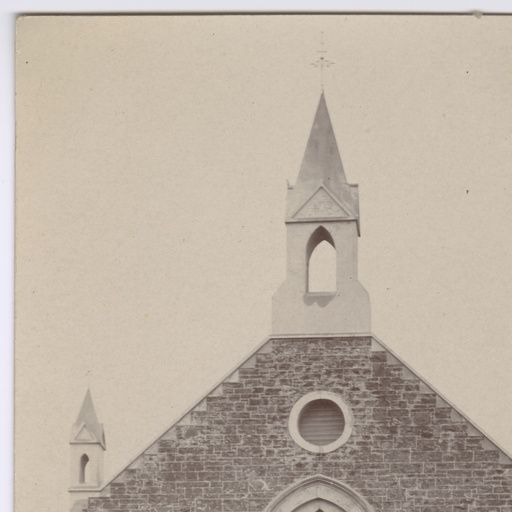 Sepia close-up of the upper gable of Morphett Vale Baptist Church — a stone spire with cross at the peak, a round porthole window in the gable, and a smaller decorative tower to the left