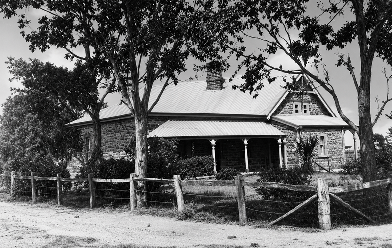 Black-and-white photograph of a stone schoolhouse with a verandah and corrugated iron roof surrounded by gum trees, behind a post-and-rail fence on a dirt road