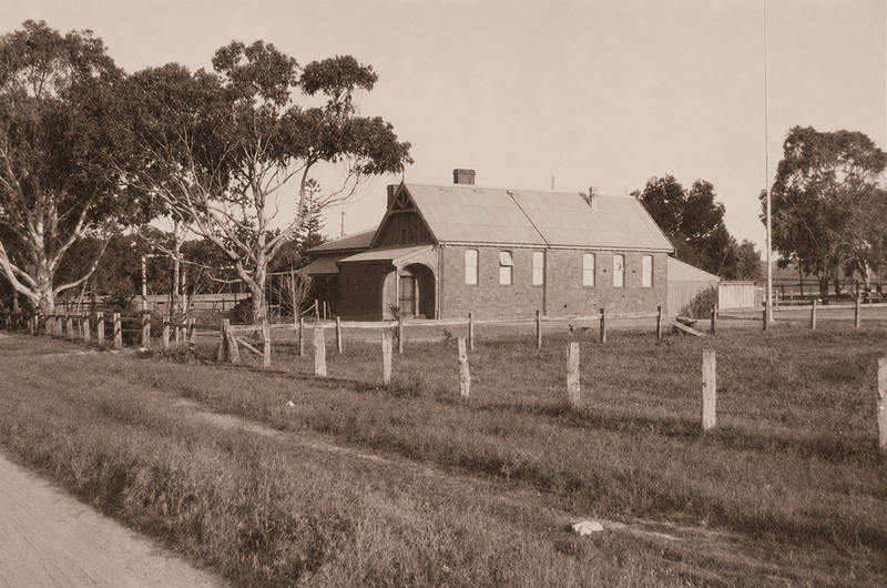 Sepia photograph of a single-storey brick schoolhouse with a corrugated iron roof, Gothic-arched windows, and a flag pole, in a fenced paddock with gum trees behind