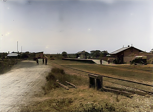 Early colour photograph of Morphett Vale station yard showing the level crossing, station building, and goods shed beside the tracks, with two figures on the platform