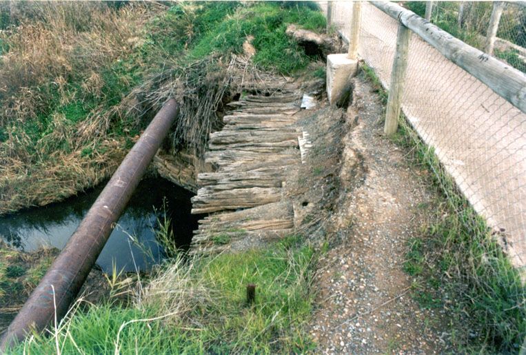Colour photograph looking along a low bridge deck of weathered grey timber logs laid side by side over a narrow creek, a rusted pipe crossing the water at left and a wire-mesh fence and gravel footpath running along the right