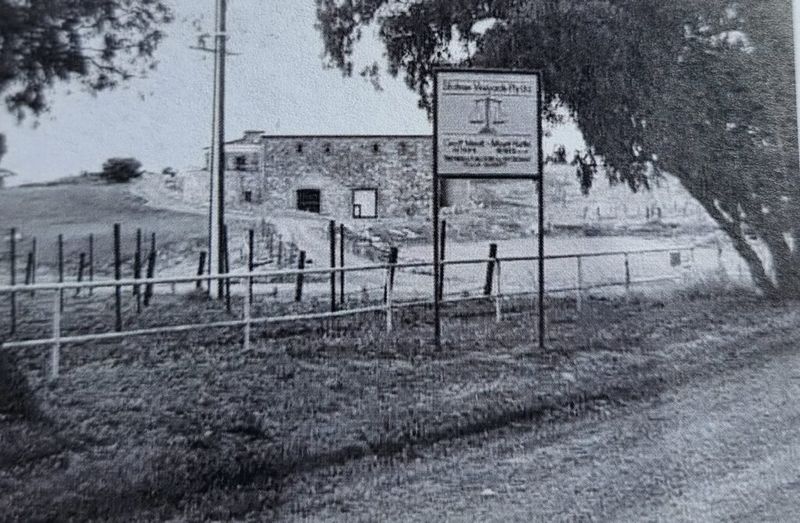 Black-and-white photograph of the Mount Hurtle Winery entrance — a two-storey stone cellar building behind a gate and winery signage, with a vine trellis in the foreground and a wooded hill behind